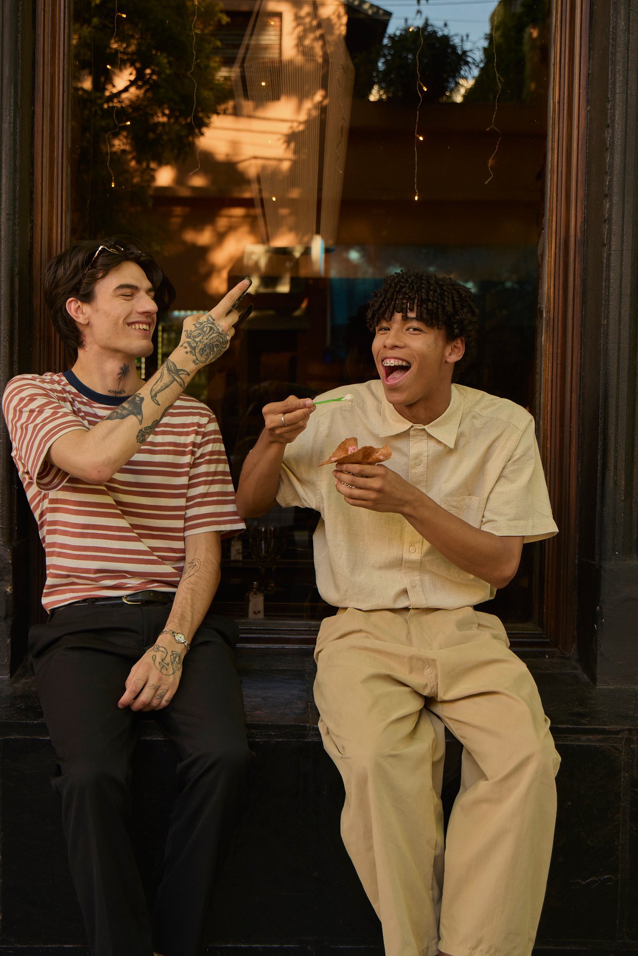 Two friends laughing on a doorstep, one feeding the other ice cream